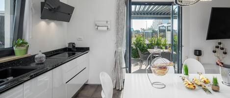 Une cuisine moderne avec un plan de travail noir, des armoires blanches et un coin repas comprenant une table et des chaises blanches. On y trouve une fenêtre avec des rideaux, un lustre et une vue sur la ville.