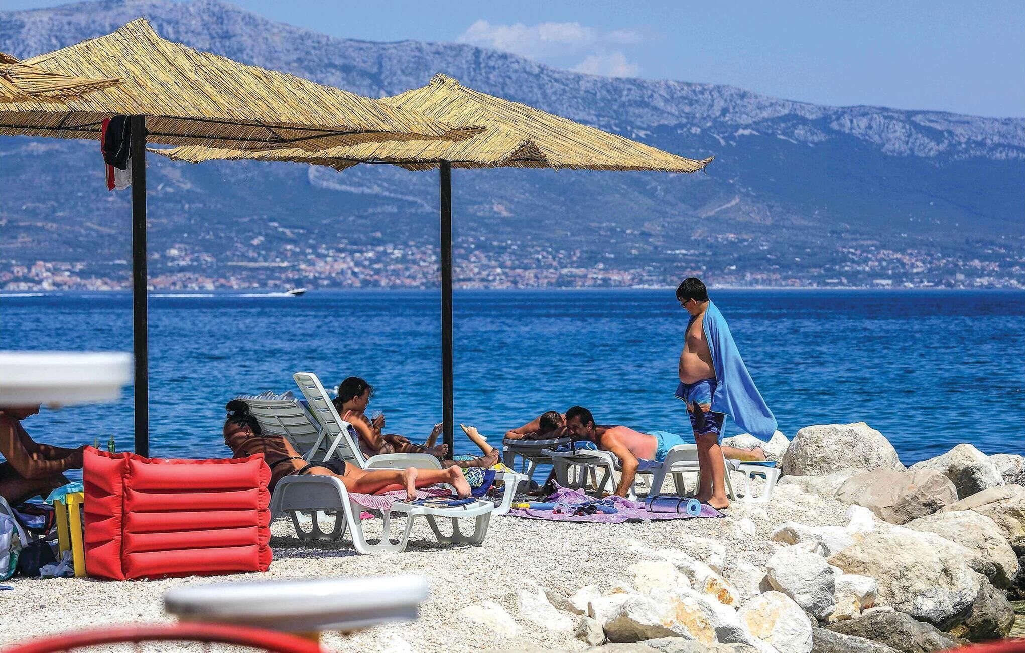 A beach with lounge chairs and umbrellas, rocky shoreline, and a view of the sea and distant mountains.
