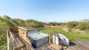 Een houten terras met een jacuzzi en barbecueplek, met uitzicht op een grasrijk landschap en de zee.