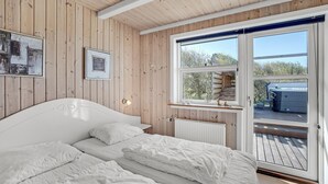 Een slaapkamer met een houten plafond, een groot bed, een raam met uitzicht op een terras en bomen, en een ingelijst kunstwerk aan de muur.