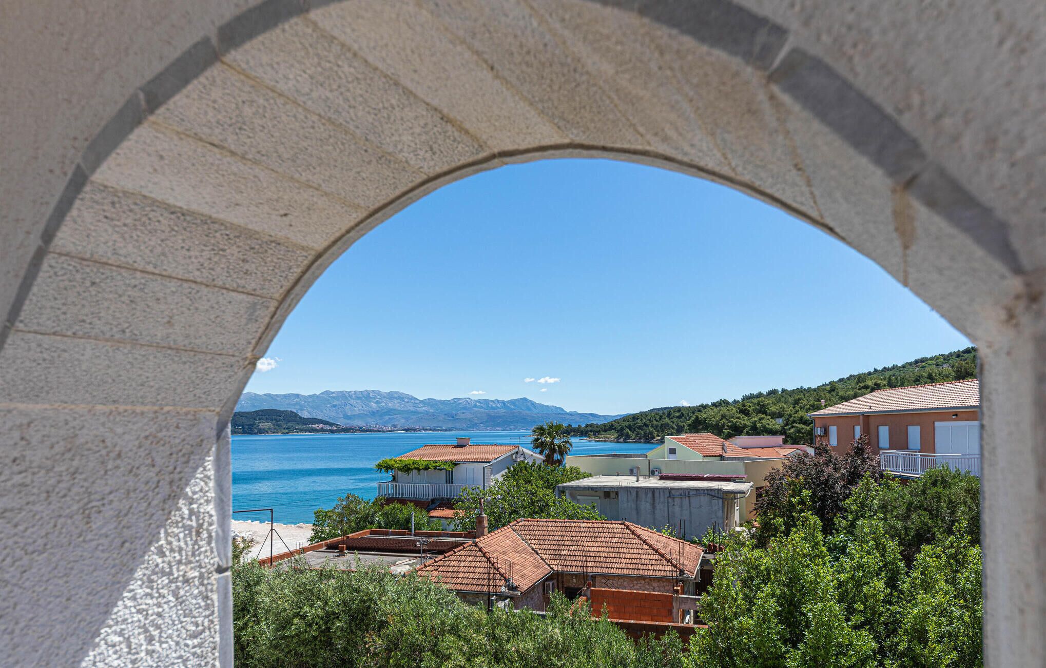 Vista costera a través de un arco, con mar azul cristalino, montañas al fondo y edificios residenciales con techos de tejas rojas.