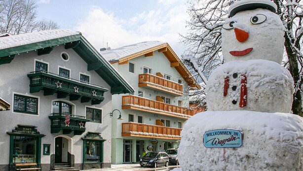 Un bonhomme de neige, devant un immeuble doté de balcons, tient une pancarte sur laquelle on peut lire : « WILLKOMMEN IN Wagrain ». Une autre pancarte, fixée à l’immeuble, indique : « FLASH PENSION ».