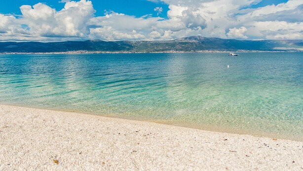 Una playa con agua turquesa cristalina, una orilla arenosa y montañas lejanas bajo un cielo parcialmente nublado.