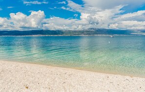 Una playa con agua turquesa cristalina, una orilla arenosa y montañas lejanas bajo un cielo parcialmente nublado.