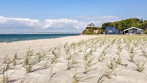 Ein Strand mit einem modernen Haus, einer Brücke im Hintergrund und spärlicher Vegetation.