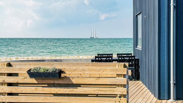 Eine Holzterrasse mit einer Bank und einem Blumenkasten, mit Blick auf ein ruhiges Meer, in der Ferne ein Schiff.