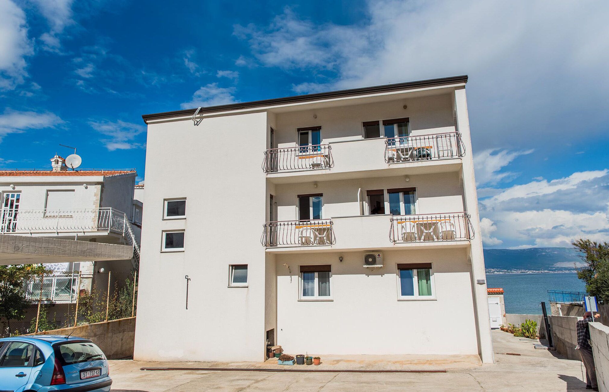 A multi-story building with balconies, a car parked in front, and a view of the sea.