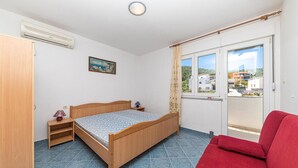 A bedroom with a bed, a red sofa, a window with a view of buildings, a painting on the wall, and a wooden door. - 1 bedroom beach front apartment in Slatine (Slatine)