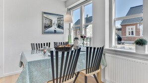 A dining area with a table set for four, a painting on the wall, and a view of a residential building through large windows. - Gorgeous home in Tranekær with kitchen (Tranekær)