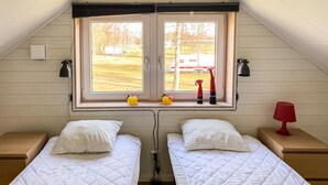 A room with two beds, a window with a view of a truck and trees, a small table with a lamp, and a red lamp on a bedside table.