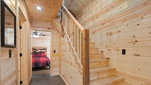 Un intérieur en bois avec un escalier, un lit recouvert d’une couverture à carreaux rouges et noirs, et un ventilateur de plafond.