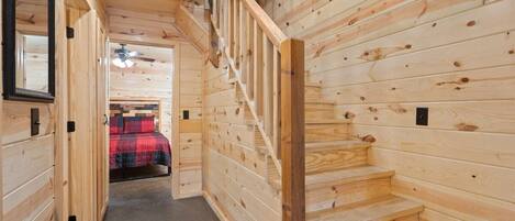 A wooden interior with a staircase, a bed with a red and black checkered blanket, and a ceiling fan.