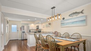A dining area with a wooden table and chairs, a kitchen in the background, and a fish-themed wall art. - Stunning Top-Floor Oceanfront Condo! Pools & New Step in Shower and More! (Nags Head)