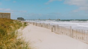 A beach with a wooden walkway, a building, and a pier extending into the ocean. - Stunning Top-Floor Oceanfront Condo! Pools & New Step in Shower and More! (Nags Head)