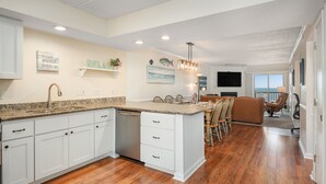 A spacious kitchen with white cabinetry, a granite countertop, and a dining area with a brown leather couch and wooden chairs. - Stunning Top-Floor Oceanfront Condo! Pools & New Step in Shower and More! (Nags Head)