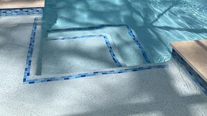 Une piscine aux eaux cristallines, bordée de carreaux bleus, entourée d’une zone abritée avec un toit en métal et des murs en verre, le tout se fondant dans un décor de verdure.