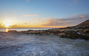 Un paysage côtier avec un coucher de soleil sur l’eau, des dunes herbeuses et un ciel dégagé.