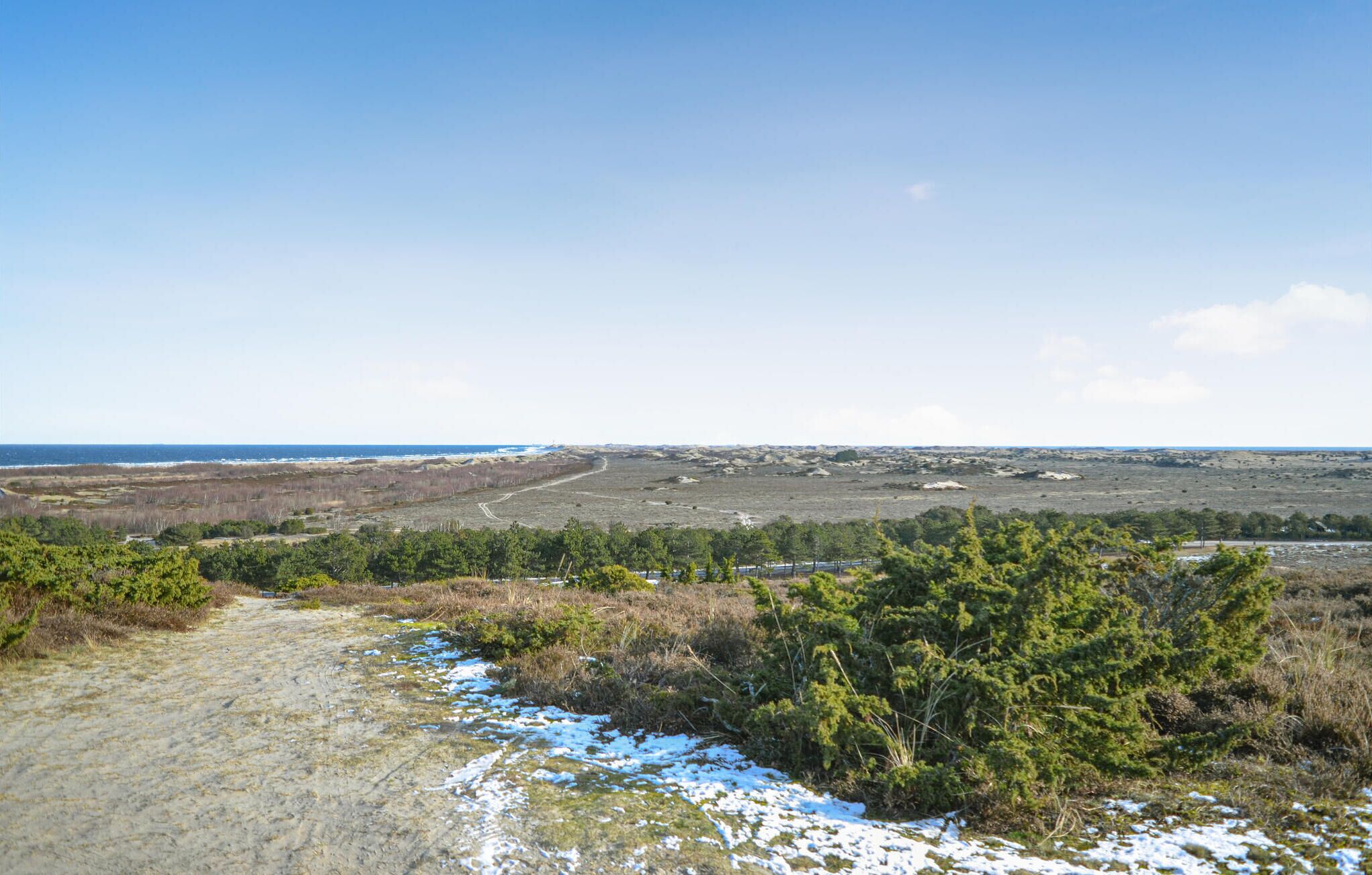 Een kustlandschap met een onverharde pad dat naar een strand leidt, omgeven door schaarse begroeiing en een heldere lucht.