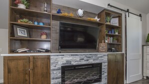 Un salon moderne doté d’un grand téléviseur à écran plat fixé au-dessus d’une cheminée, d’étagères en bois ornées d’objets décoratifs et d’une porte coulissante située à droite.