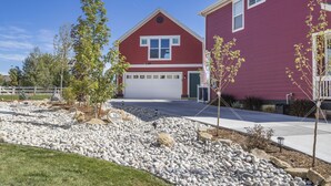 Une maison rouge à deux étages, avec une porte de garage blanche, entourée d’un jardin aménagé avec des rochers et de jeunes arbres.