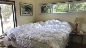 Un lit avec une literie blanche, une fenêtre donnant sur des arbres et un tableau encadré au mur.