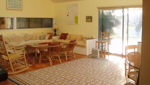 Un salon avec un canapé, des fauteuils en osier, un tapis à motifs et une grande fenêtre donnant sur un beau paysage.