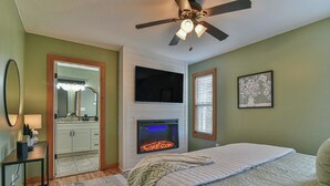 Une chambre à coucher comprenant un lit, une cheminée, un ventilateur de plafond, un miroir et une fenêtre avec des stores.