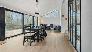 Uma ampla sala de jantar com uma mesa comprida e cadeiras, janelas grandes com vista para o exterior e um design de teto moderno.