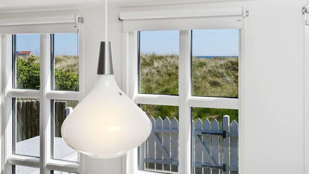Een witte hanglamp hangt voor een raam met uitzicht op een witte schutting en grasduinen.