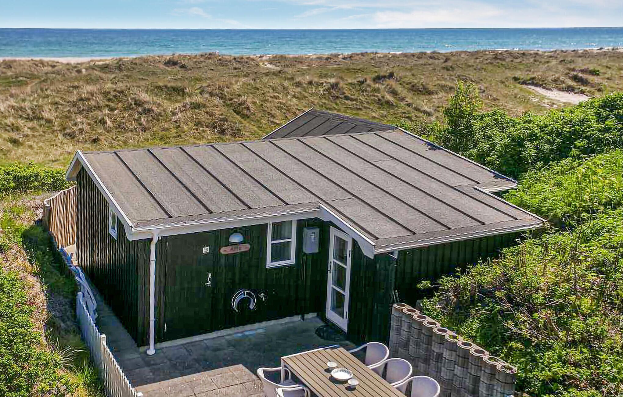 A small wooden cabin with a sloped roof, a patio with a table and chairs, and a view of the beach and ocean.