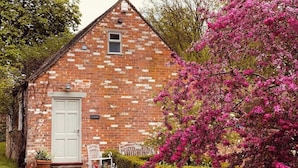 A brick building with a white door, a small window, and a garden with pink flowers and a hedge. - Refurbished Coach House in rural setting. (Waddesdon)