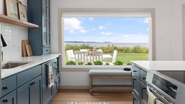 A kitchen with a large window, wooden cabinets, a bench, and a view of the outdoors. - Beach Front Cottage in the Sun on Whidbey. (Greenbank)