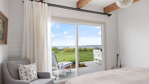 A bedroom with a large bed, a grey armchair, a white chair, and a view of a grassy yard through a sliding glass door. - Beach Front Cottage in the Sun on Whidbey. (Greenbank)