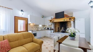 Moderni keittiö, jossa on valkoiset kaapit, sisäänrakennettu uuni ja takka. Keittiössä on myös sohva sekä ruokapöytä tuoleineen.