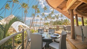 Un espacio de comedor al aire libre con mobiliario de mimbre, una terraza de madera y vistas a palmeras y al océano.