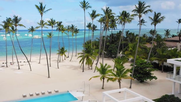 Vista de la playa con palmeras, piscina y un edificio blanco.
