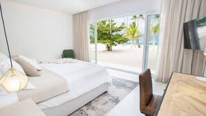 Una habitación de hotel con una cama, un escritorio, una silla, un televisor y un ventanal con vistas a palmeras y una playa.