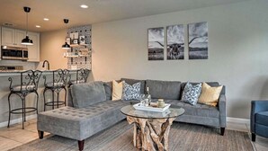 A modern living room with a grey sectional sofa, a round coffee table with a tree stump base, and a kitchen in the background.