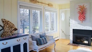 A cozy living room with a white fireplace, a bench with cushions, and a large window with blinds.