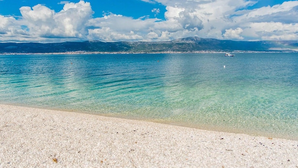 Ein Strand mit klarem türkisblauem Wasser, einem sandigen Ufer und fernen Bergen unter einer teils bewölkten Himmel.