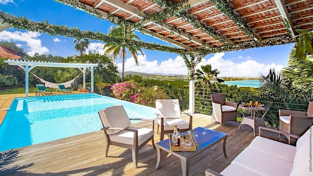A poolside area with a wooden deck, lounge chairs, a table with drinks, and a view of the sea and mountains.