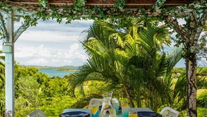 An outdoor dining area with a table set for four, surrounded by lush greenery and a view of the sea.
