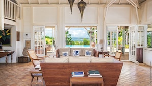 A spacious living area with a large sofa, wooden coffee table, and two hanging lamps. There are open doors leading to a balcony with a view of the ocean.