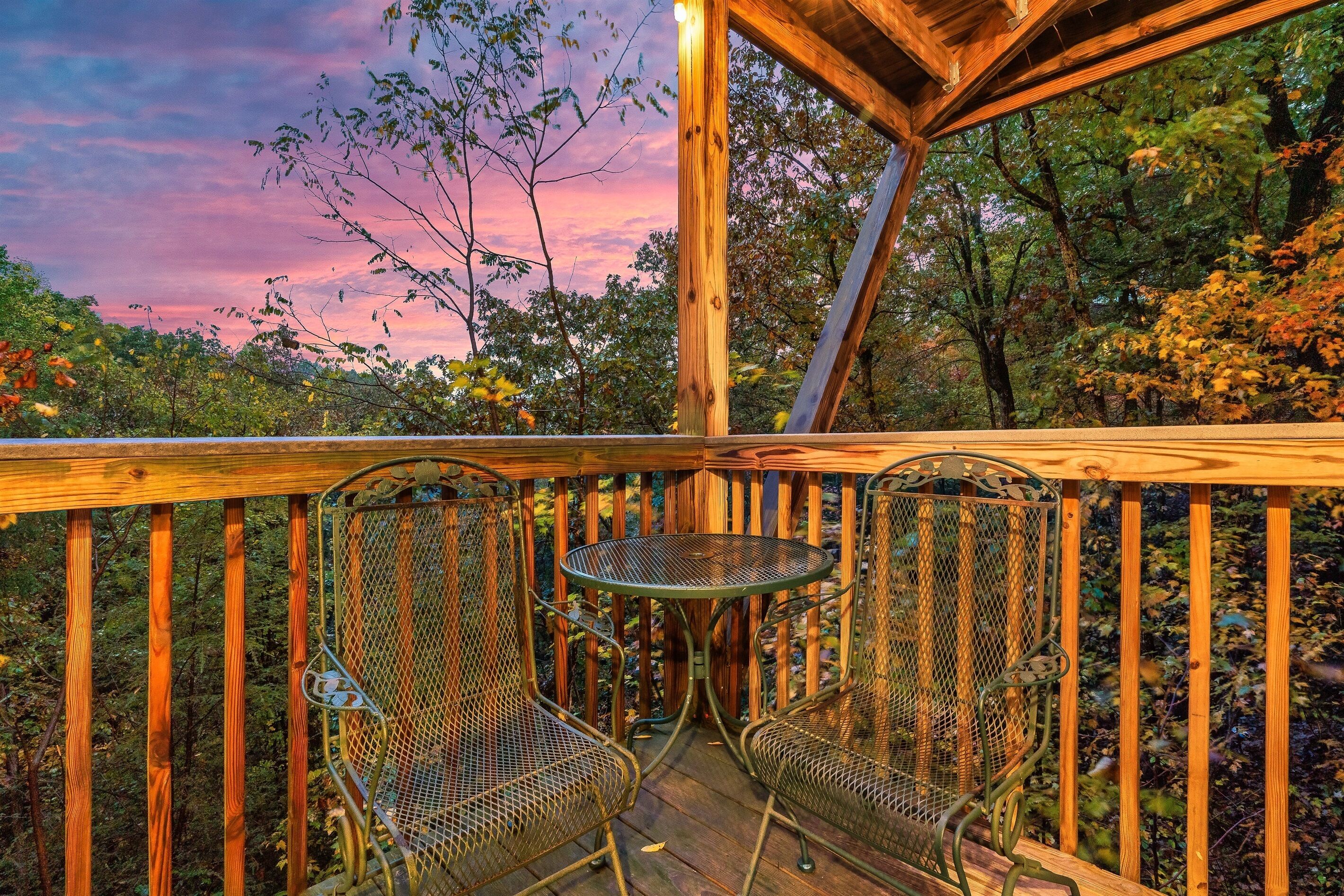 Une terrasse en bois avec des chaises en métal et une petite table, entourée d’arbres et d’une rambarde en bois.