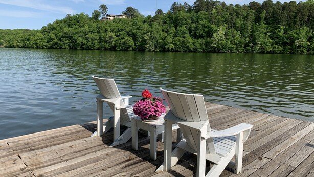 Une jetée en bois avec deux chaises Adirondack blanches et un pot de fleurs contenant des fleurs roses.