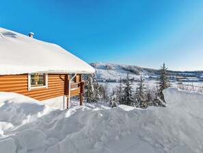 Une cabane en bois, dont le toit est recouvert de neige, entourée d’un paysage enneigé et surplombée par un ciel bleu dégagé.
