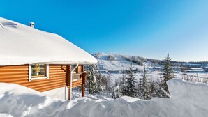 Eine Holzhütte mit verschneitem Dach, umgeben von Schnee und Bäumen, mit Blick auf eine verschneite Landschaft.