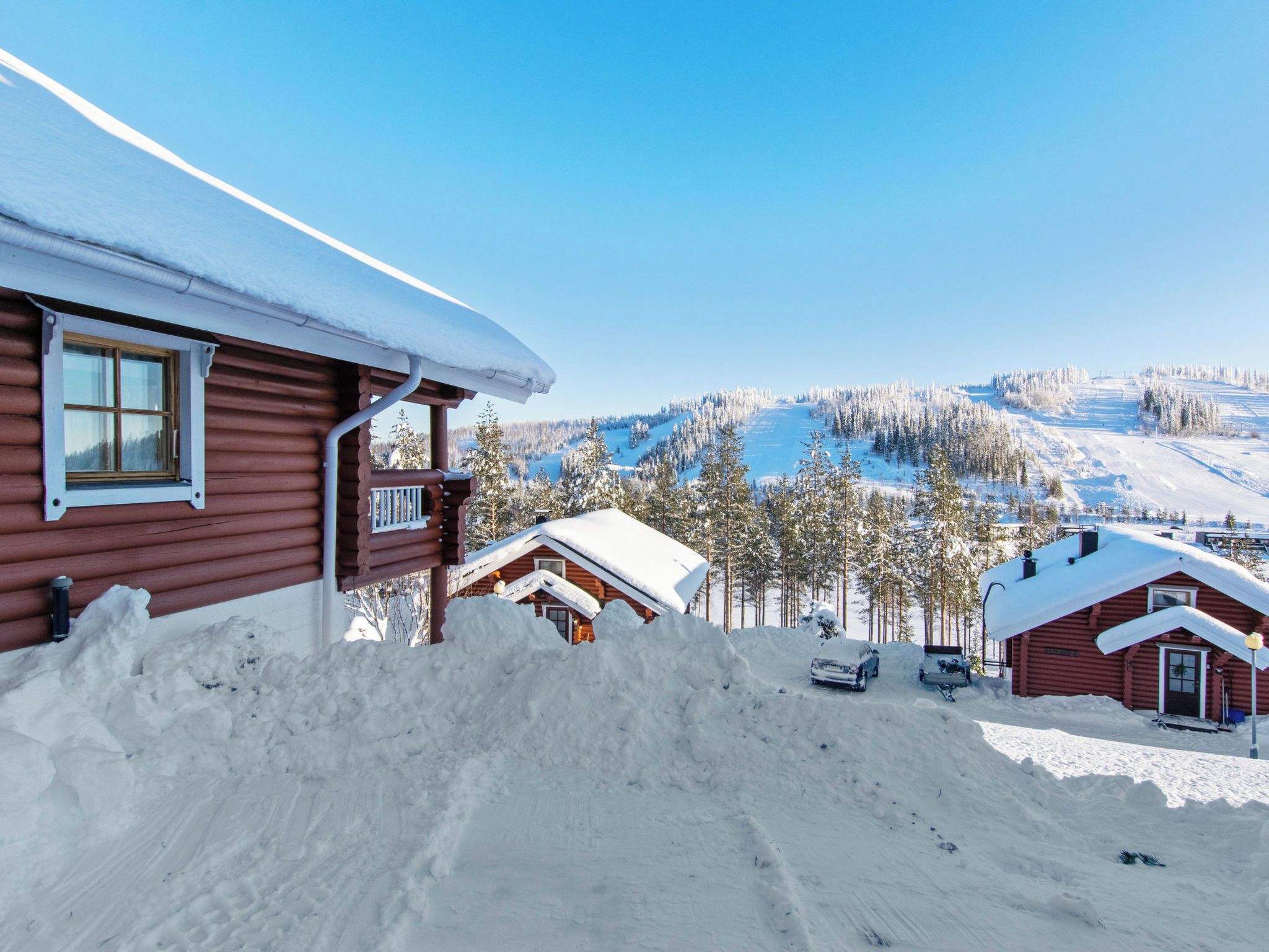 A cabin with a snow-covered roof, a wooden exterior, and a view of a snowy landscape with trees and a clear sky.