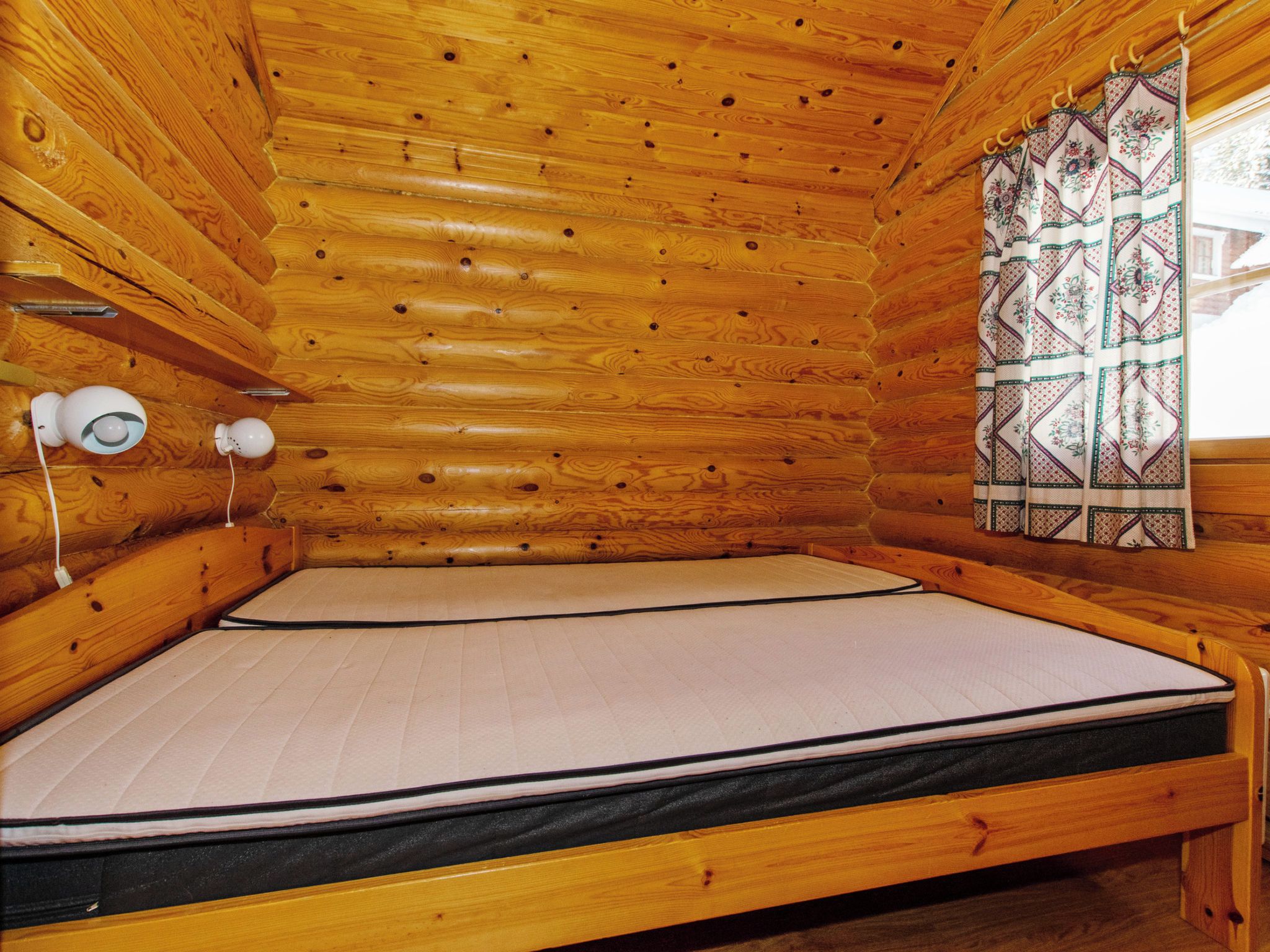 A wooden cabin room with a single bed, a window with patterned curtains, and a wall-mounted light fixture.