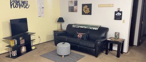 A living room with a black leather sofa, a TV, a coffee table, and a rug. There are wall decorations and a sign that says "HOME SWEET HOME".